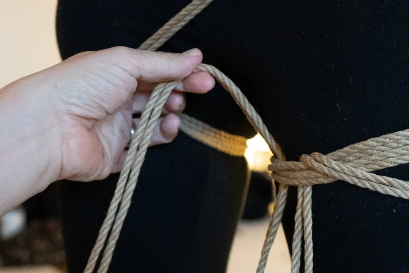 Shibari-Anleitung: Hand führt eine Seilschlaufe an der Oberschenkelfesselung, Hanfseil auf schwarzer Kleidung